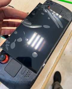 Técnico sosteniendo el nuevo panel de pantalla OLED con plástico protector sobre el cuerpo de la consola Steam Deck durante el proceso de cambio.